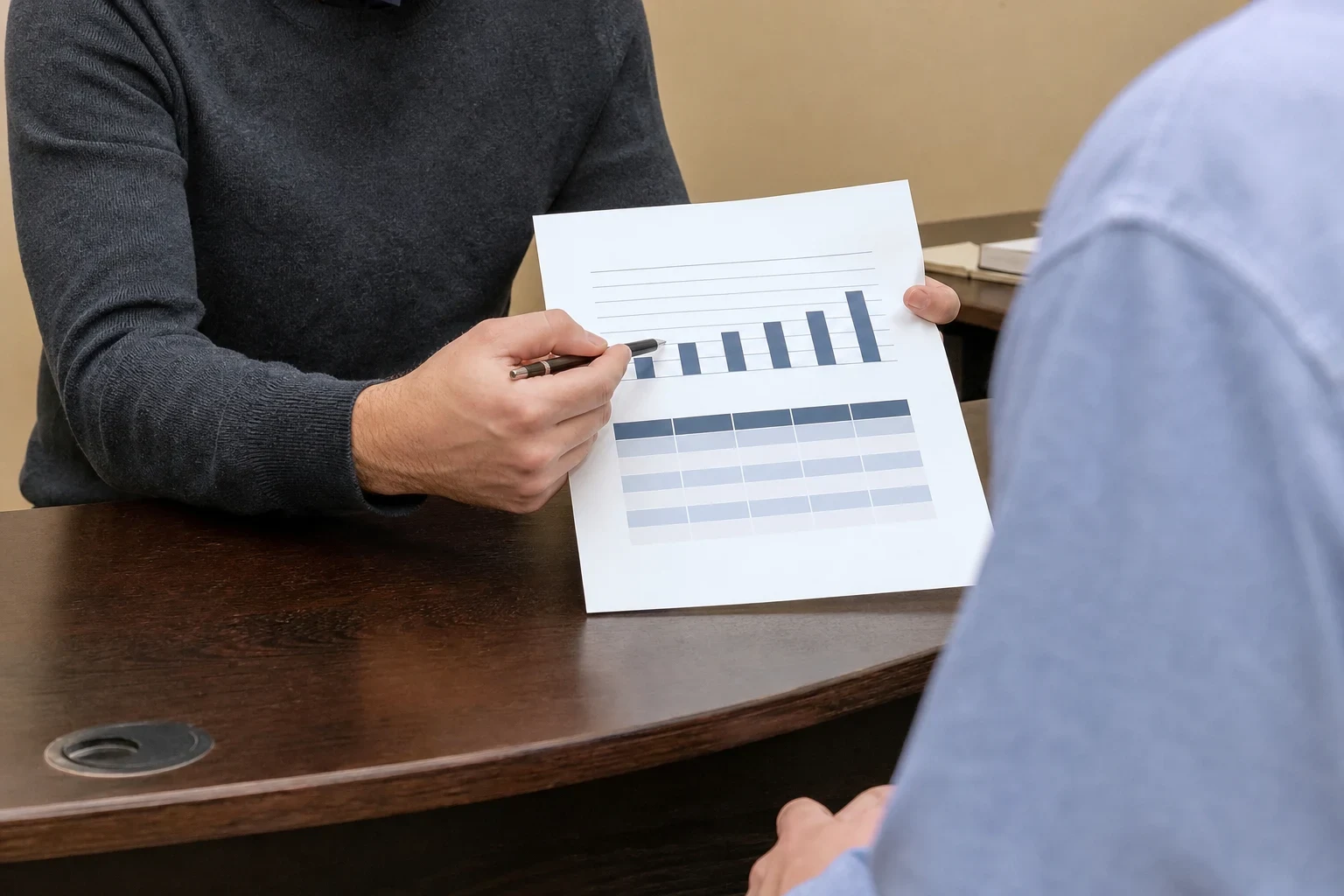 Agent in a gray sweater points a pen at a printed bar chart and table on a wooden office desk while a client in a blue shirt looks on