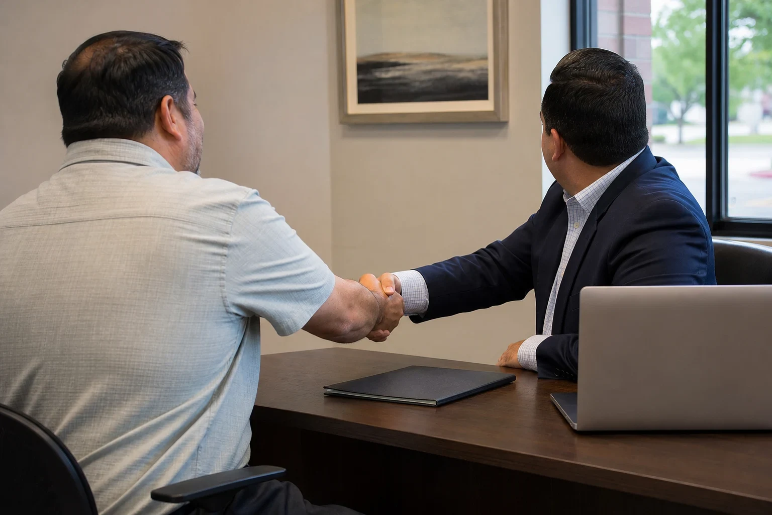 Insurance agent in a navy blazer shaking hands with a client across a wooden desk with a laptop and closed folder, framed landscape art and window behind