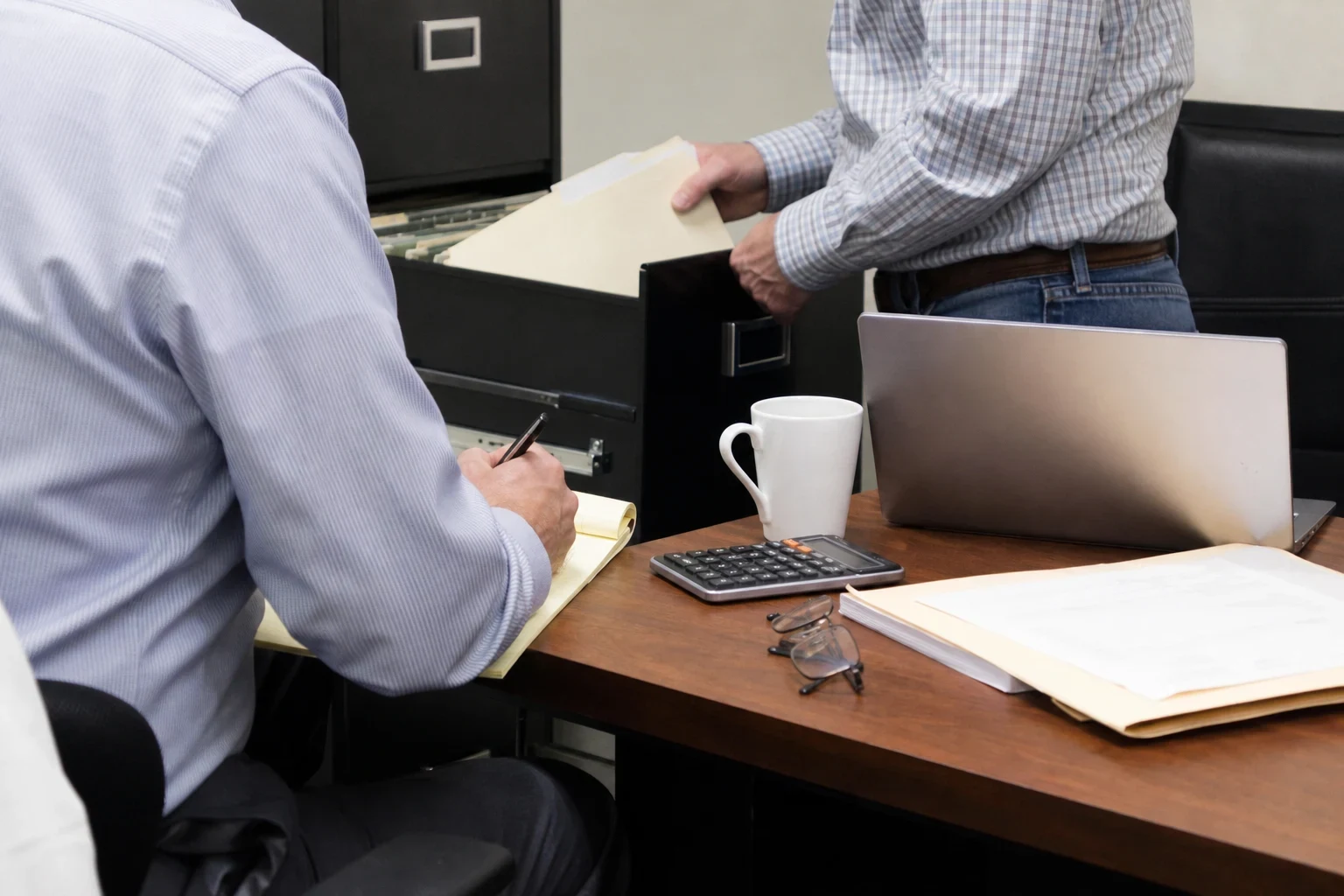 A man in a striped shirt takes notes on a yellow legal pad at a desk while another man pulls a manila folder from a black filing cabinet; calculator, mug and laptop nearby.