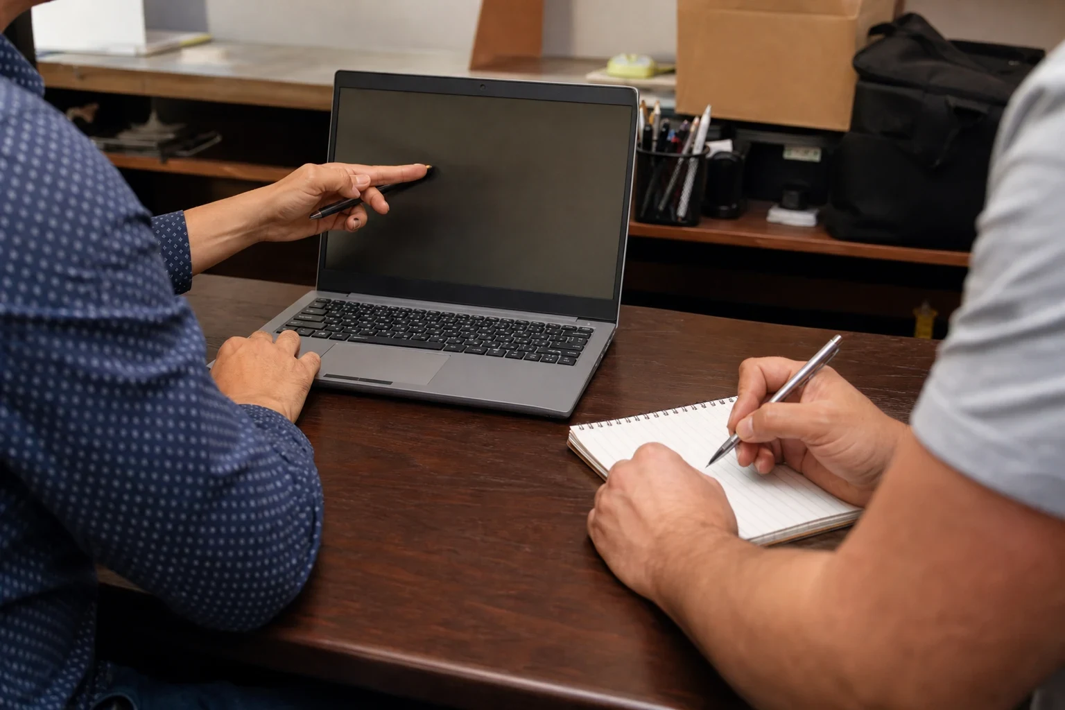 Two people at a wooden office desk, one pointing a pen at a laptop screen while the other writes notes in a spiral notebook, shelves and boxes behind